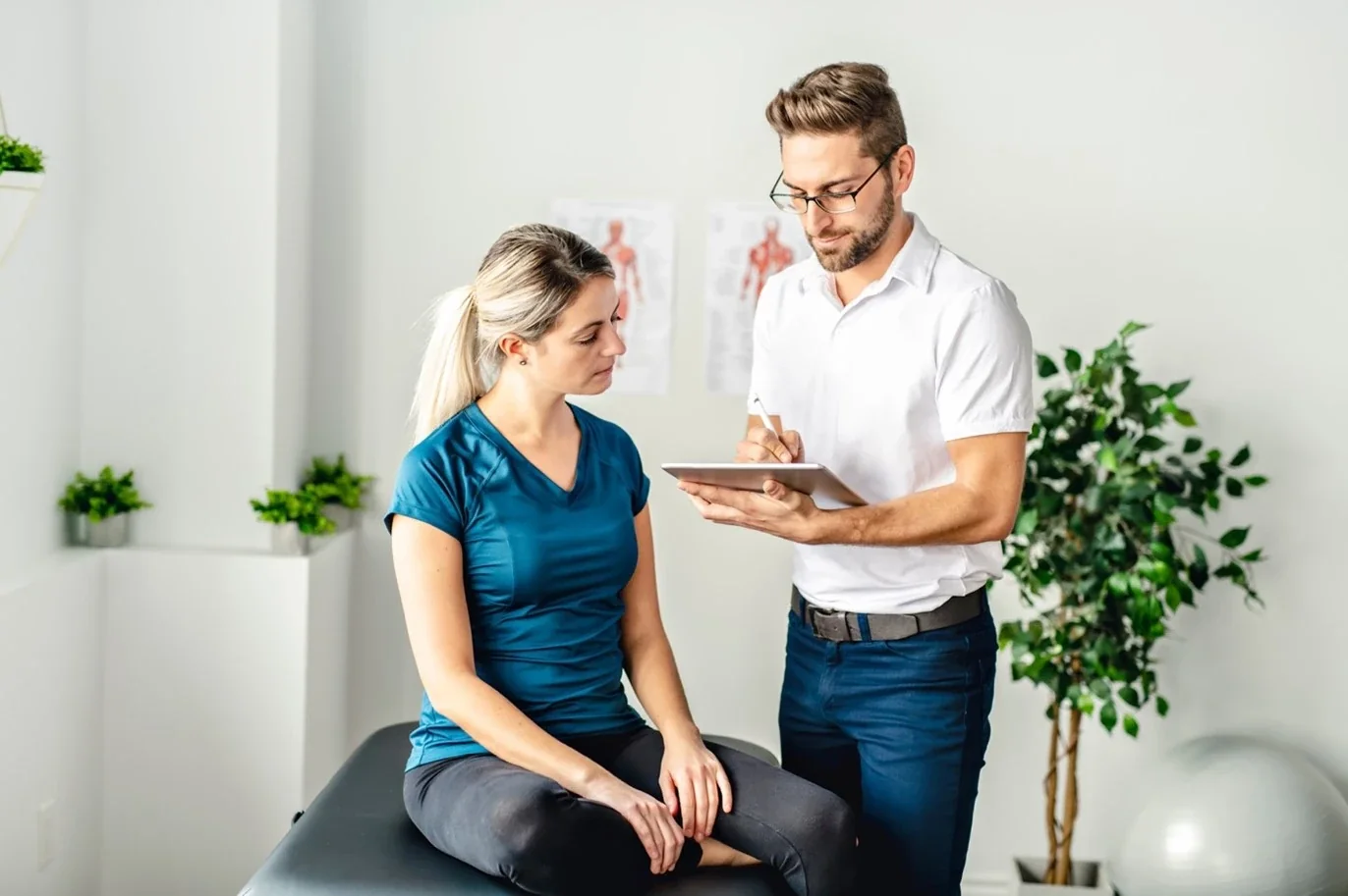 Therapist taking notes on a tablet while talking with a patient seated on a treatment table